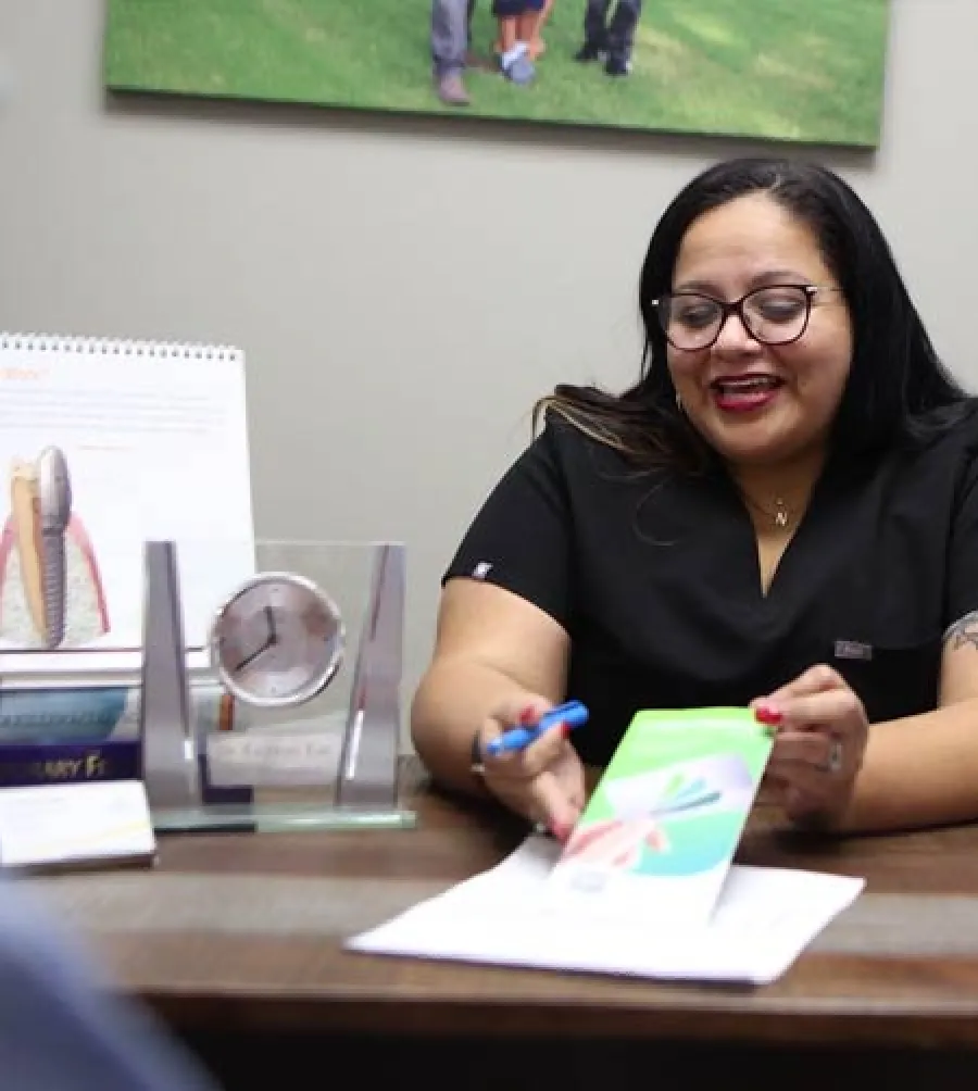 Woman in glasses explaining a document at a desk with books, clock, and a dental model in the background