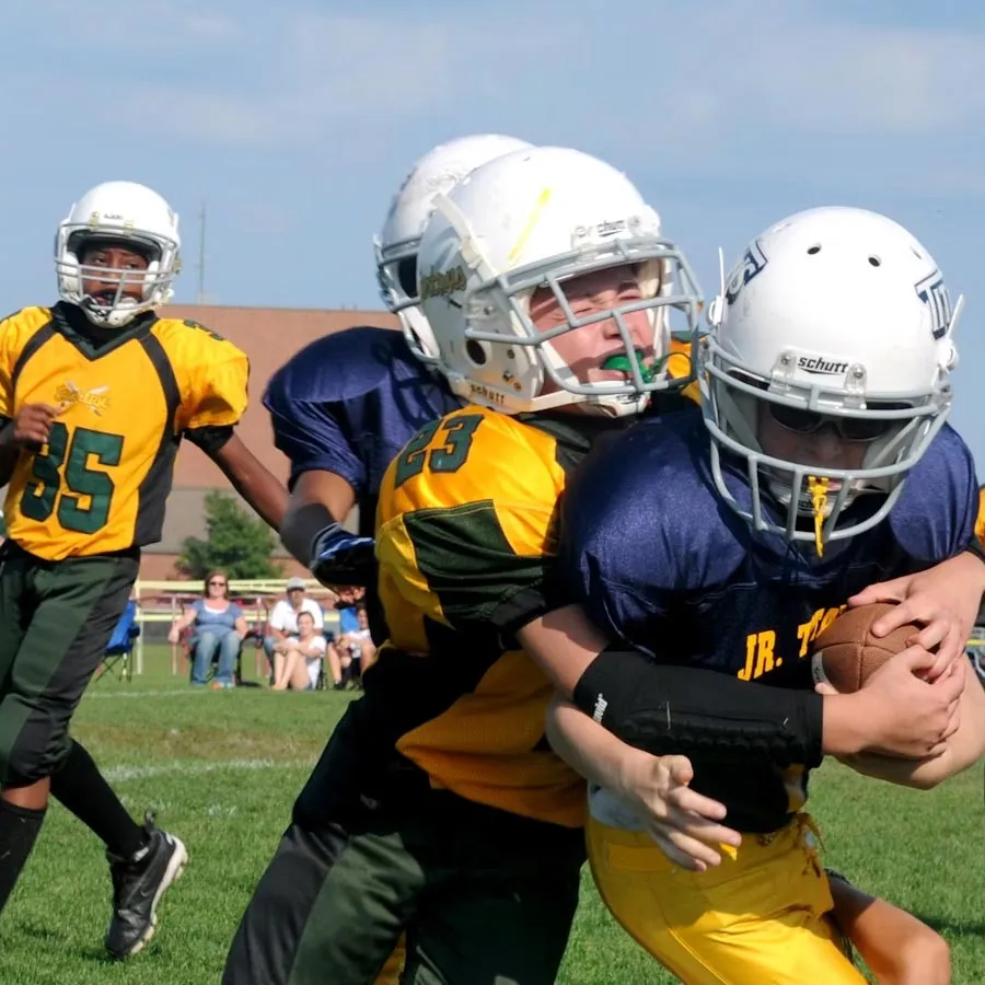 Youth football players tackling during a game with spectators in the background.