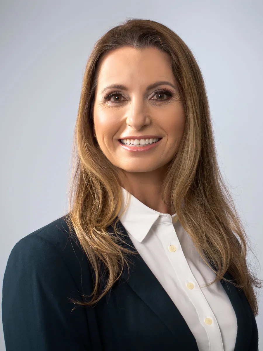 Professional woman with long brown hair wearing a white blouse and dark blazer smiling against a gray background