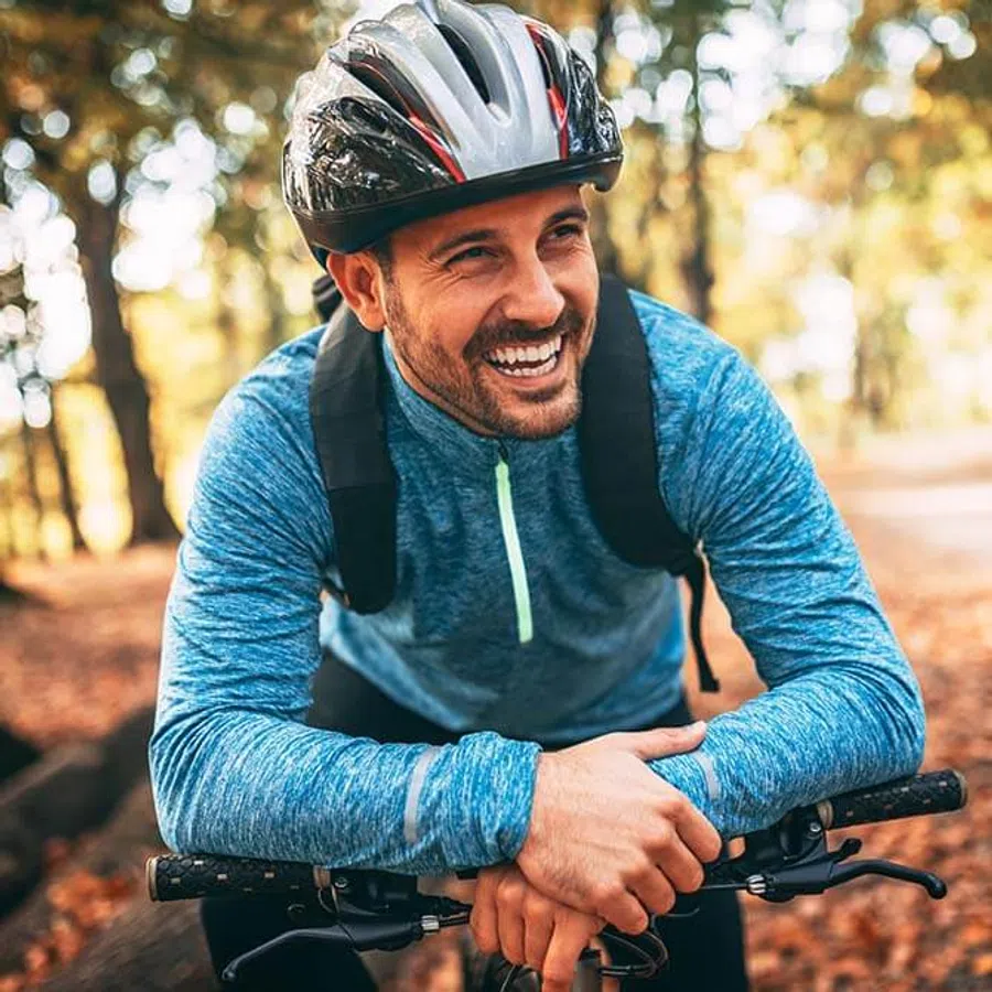 Smiling man in blue jacket and helmet resting on bike handlebars in autumn forest trail.