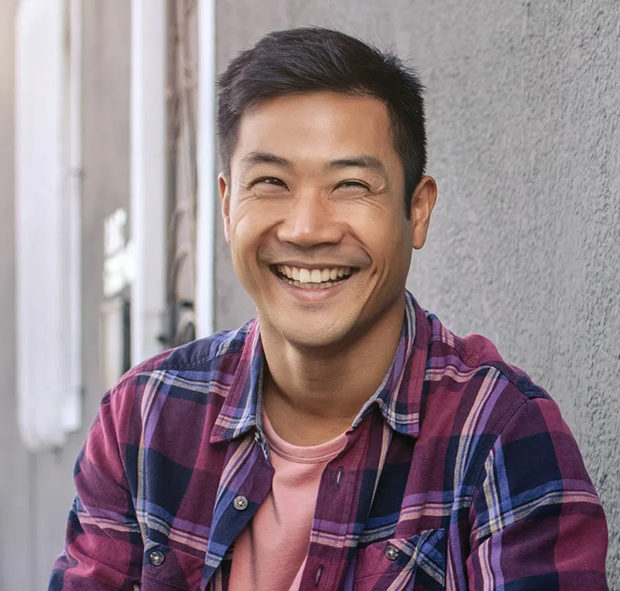 Smiling young man wearing a purple plaid shirt standing against a gray wall with natural light.