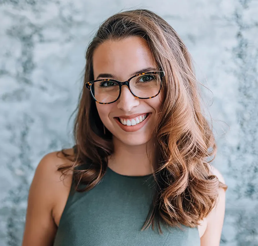 Smiling woman with long brown hair, wearing tortoiseshell glasses and a green sleeveless top, against a textured wall.