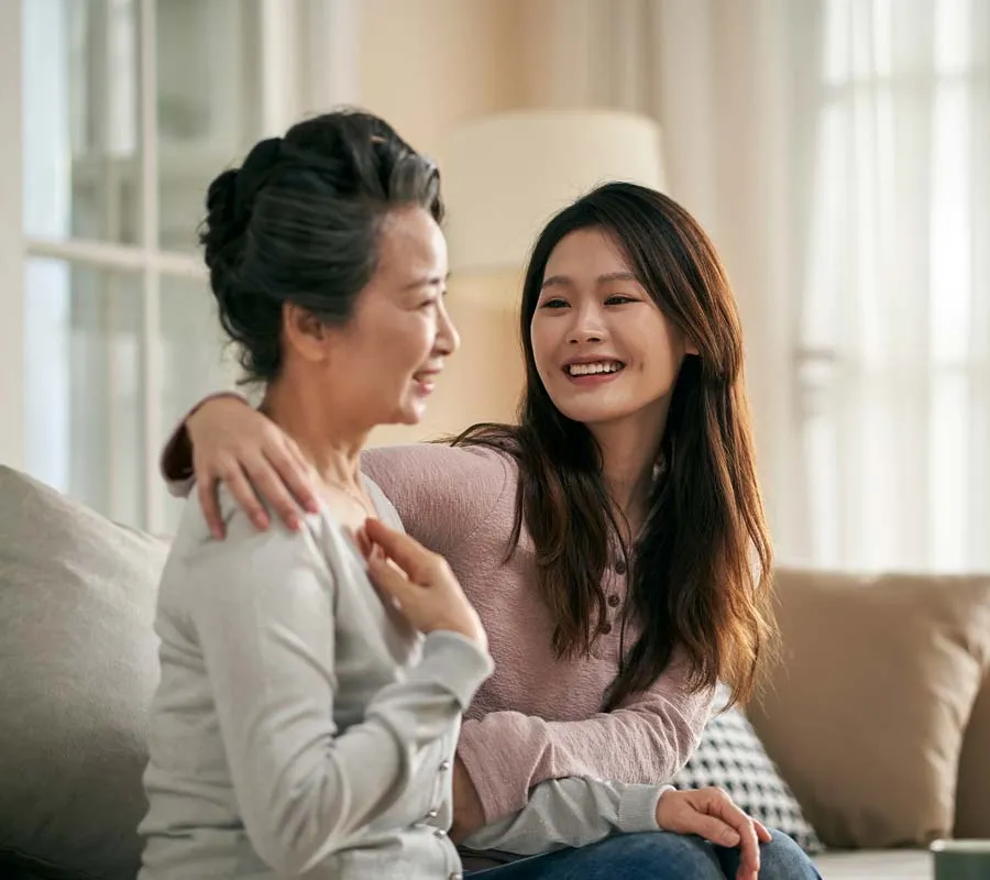 Young woman smiling and embracing older woman while sitting on a sofa in a bright living room.