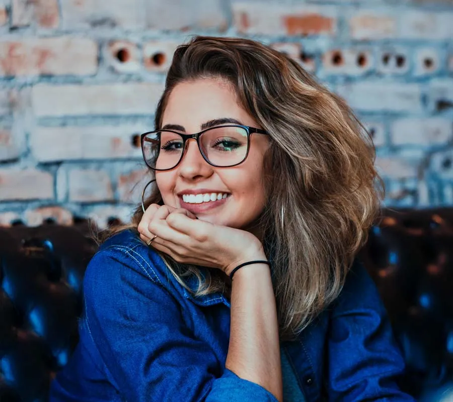 Smiling young woman with glasses and wavy hair resting her chin on hand against a brick wall background.