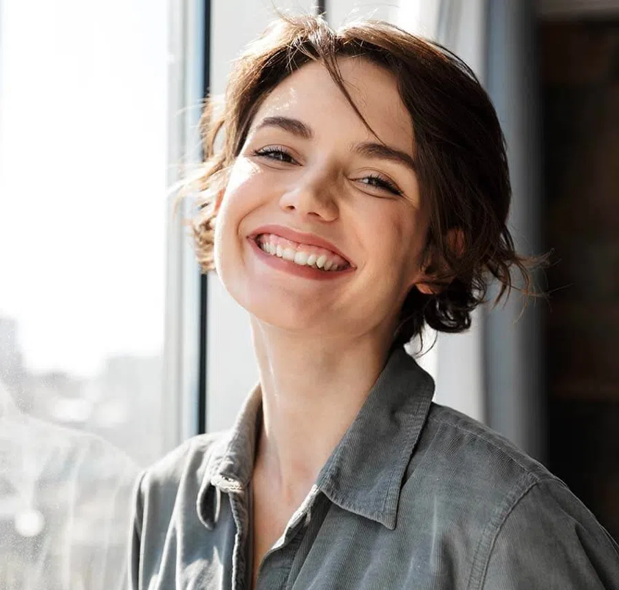 Happy young woman smiling brightly near a window in natural light wearing a gray shirt.