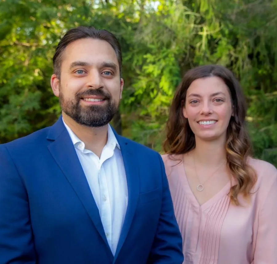 Smiling man in blue suit and woman in pink blouse standing outdoors with green trees and grass in background