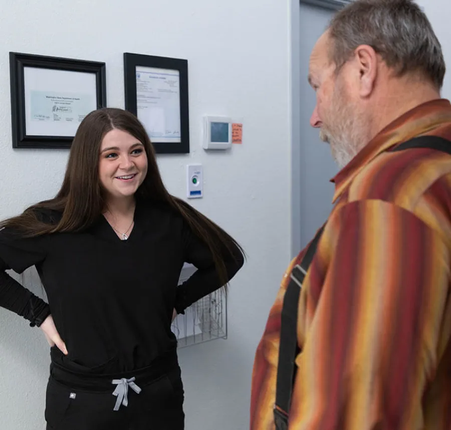 Young woman in black scrubs smiling and talking to an older man in a striped shirt in an office setting.