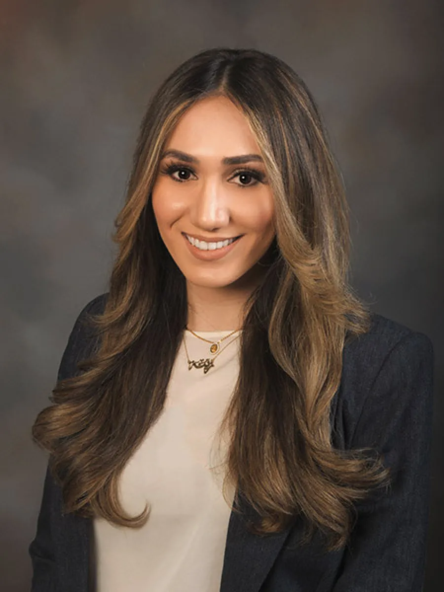 Smiling woman with long highlighted hair wearing a blazer and layered necklaces against gray background