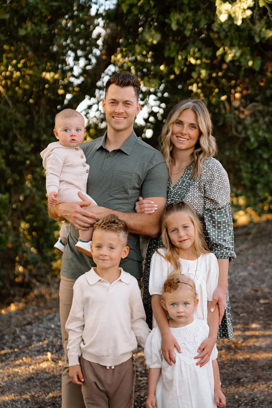 Smiling family of six with parents and four children posing outdoors in natural sunlight with green trees in background