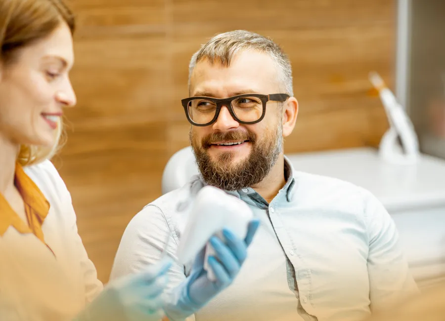Smiling male patient with glasses talking to female dentist holding a dental model in clinic.