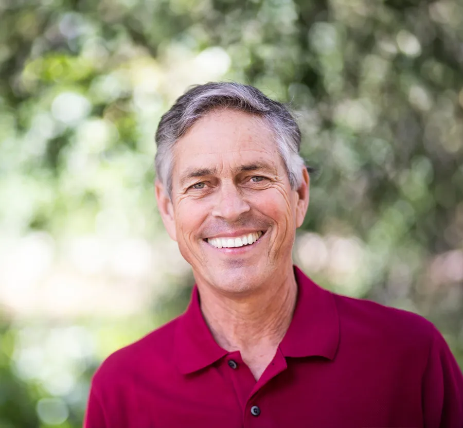 Smiling middle-aged man wearing a red polo shirt outdoors with a blurred green natural background.