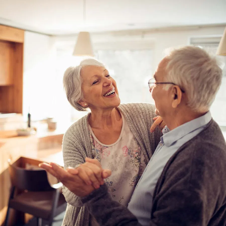Older couple smiling and holding hands while dancing affectionately in a cozy, sunlit living room.