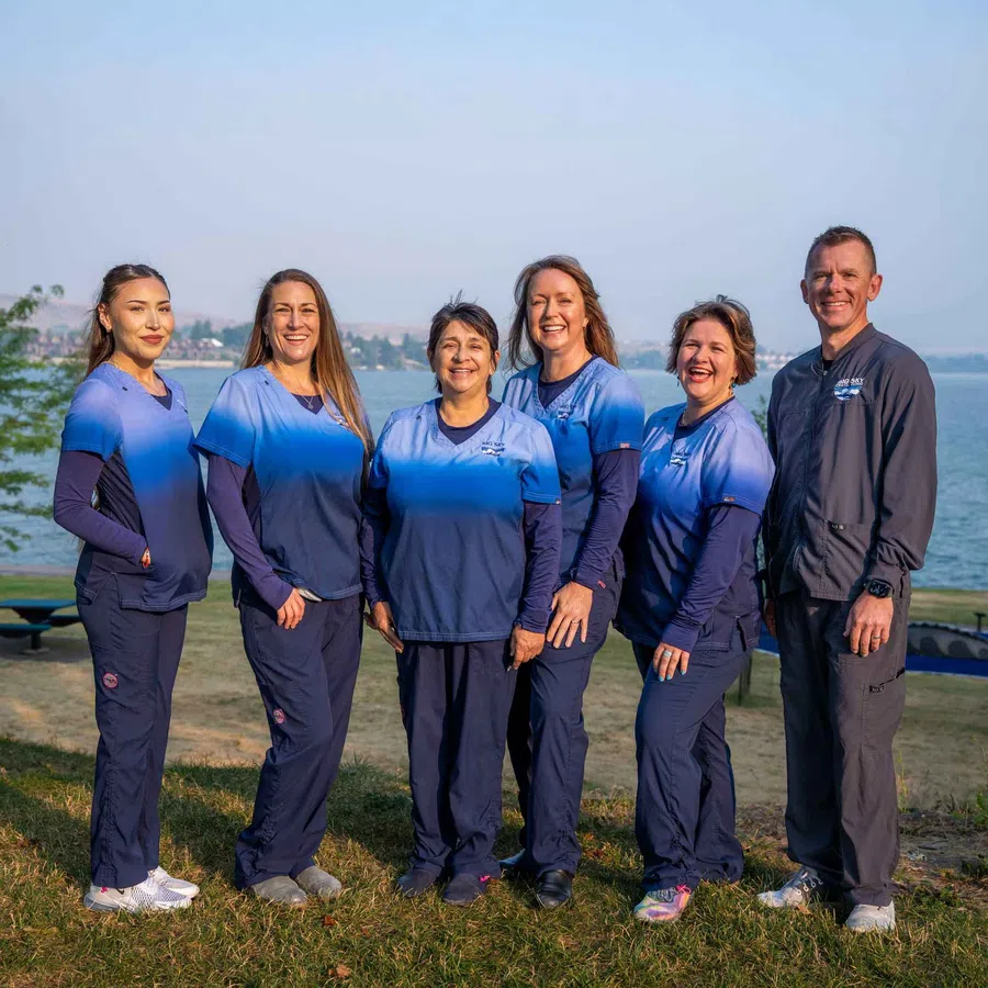 Group of six healthcare professionals in blue scrubs standing outdoors by a lake, smiling and posing for a photo.