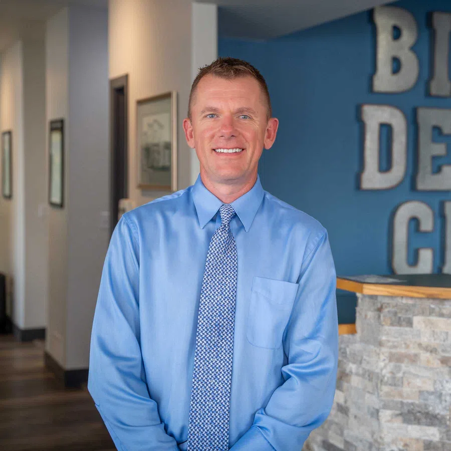 Smiling man in blue dress shirt and patterned tie standing in a modern office lobby with stone and blue wall background