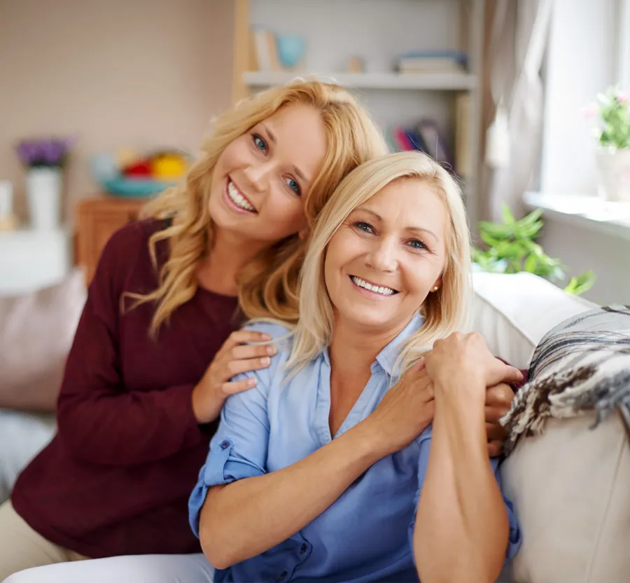 Smiling mother and adult daughter embracing on a cozy sofa in a bright living room.