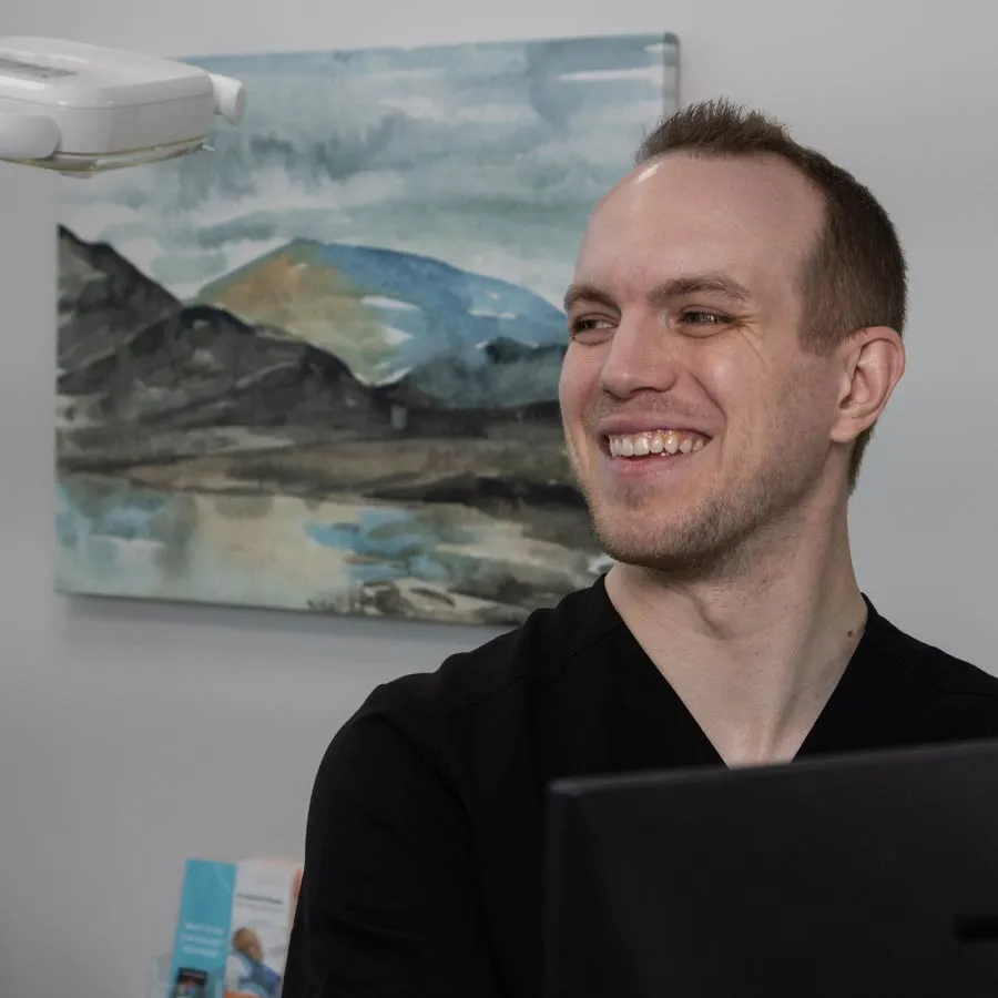 Smiling male healthcare professional in black scrubs at desk with mountain landscape painting in background