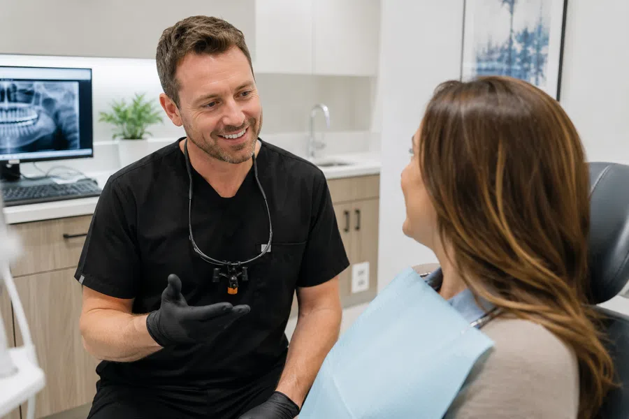 Dentist in black scrubs consulting smiling female patient in modern dental office with X-ray on screen.