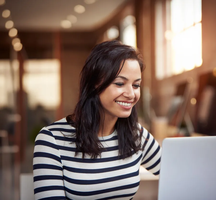 Smiling woman in striped shirt working on laptop in bright modern office with warm lighting.