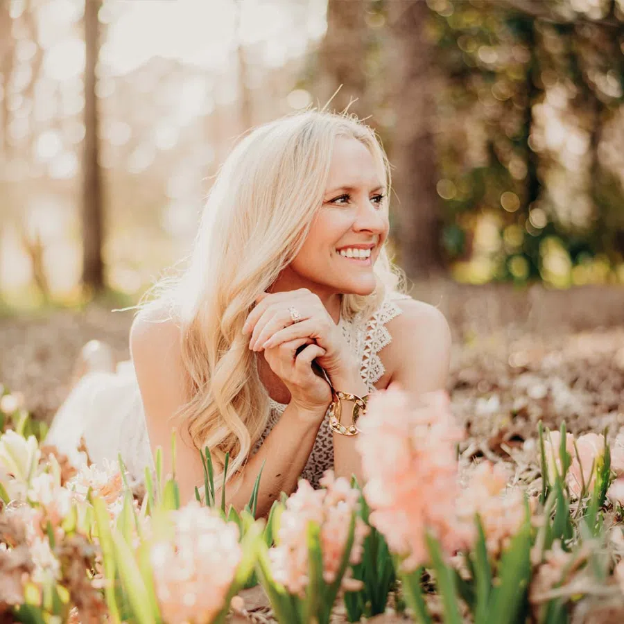 Blonde woman smiling and lying among blooming pink flowers in a sunlit forest setting.