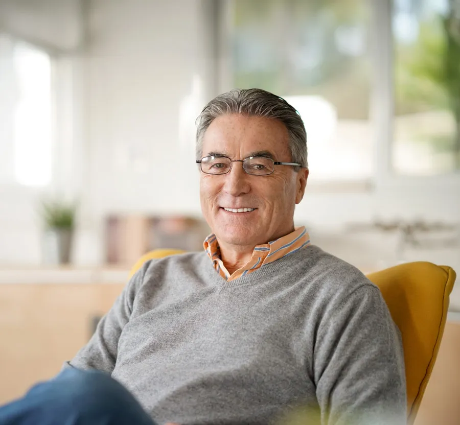 Smiling middle-aged man in glasses wearing a gray sweater sitting on a yellow chair indoors with a bright background