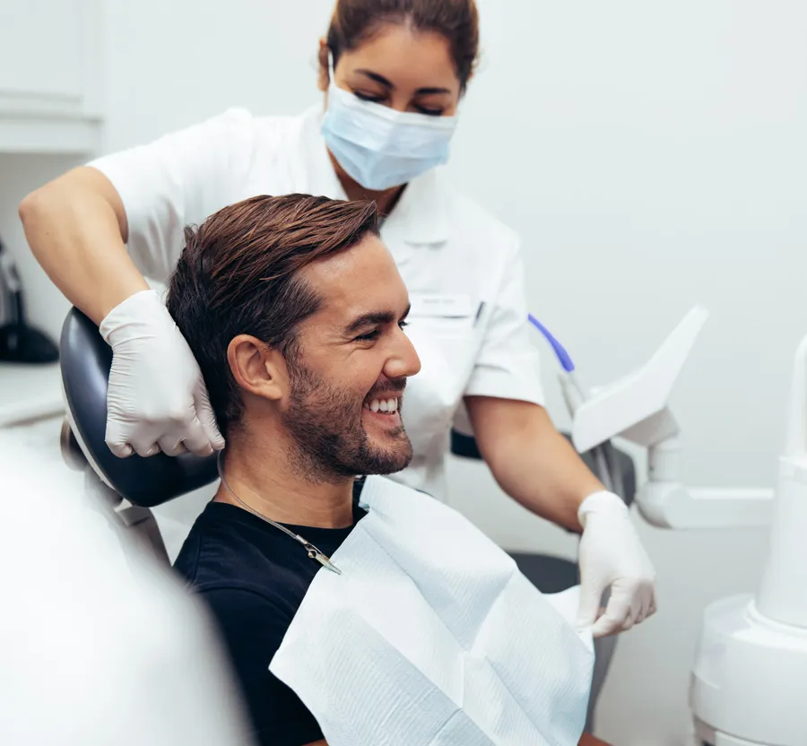 Smiling man sitting in dental chair with dental hygienist adjusting chair wearing mask and gloves