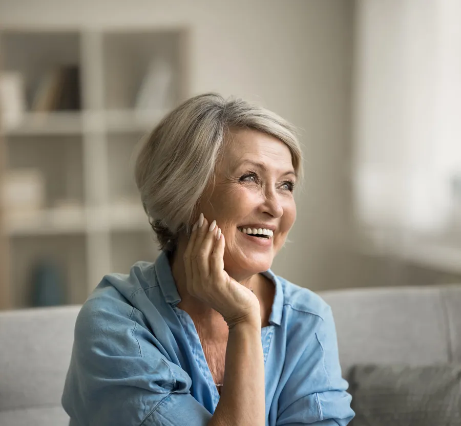 Smiling mature woman in blue shirt looking away while sitting on a couch in a bright living room