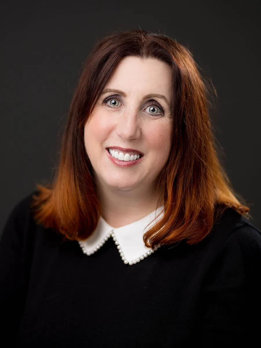 Smiling woman with red hair, pearl-adorned white collar, and black top against a dark background.