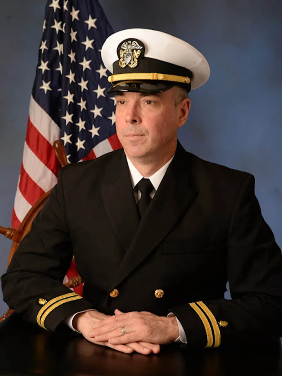 Portrait of a US Navy officer in formal uniform seated with folded hands, American flag and ship wheel in background