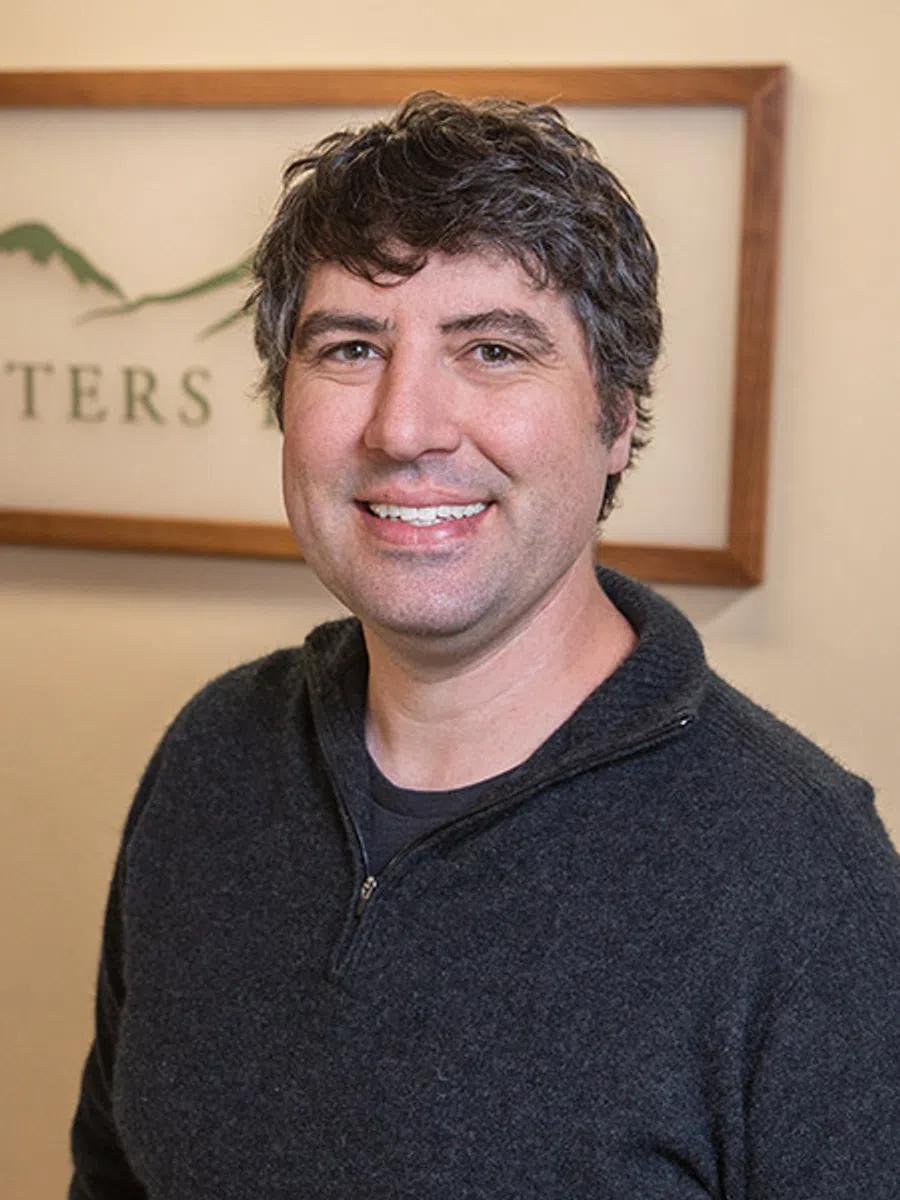 Smiling man wearing a dark sweater standing indoors with a framed mountain logo in the background