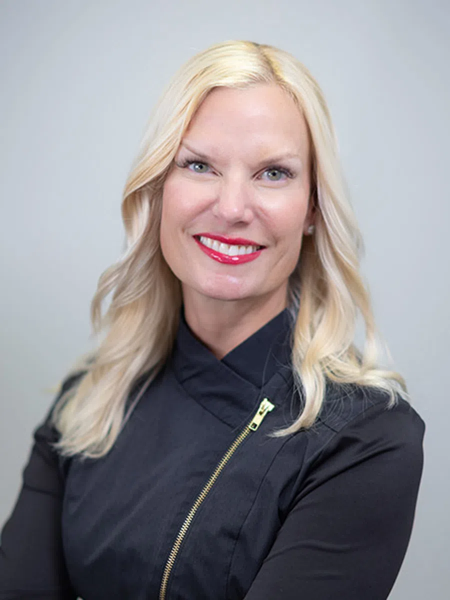 Portrait of a smiling blonde woman with red lipstick wearing a black jacket with a gold zipper against gray background