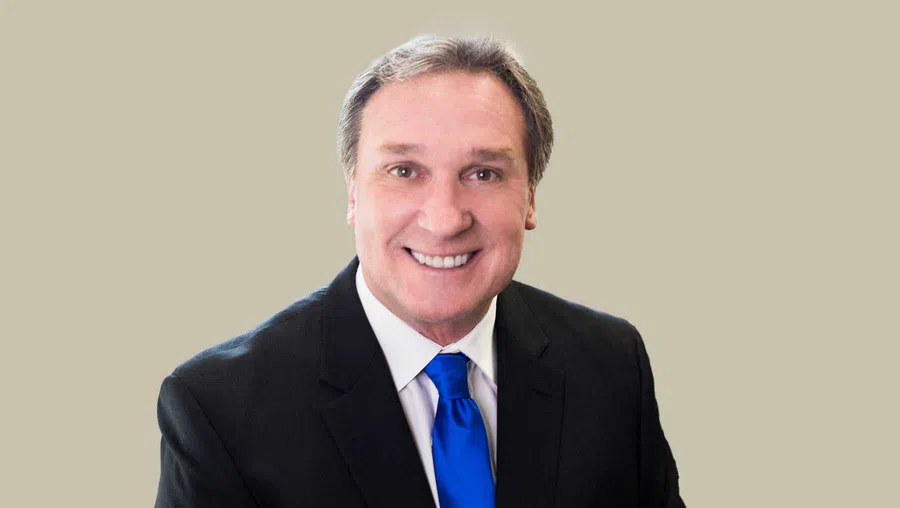 Smiling middle-aged man in a black suit with a white shirt and bright blue tie against a plain beige background