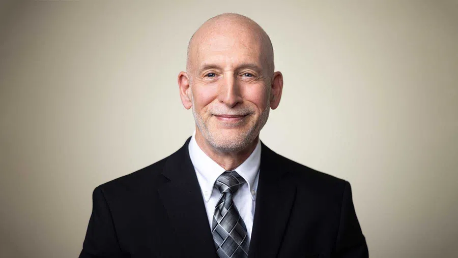 Smiling bald middle-aged man in a black suit, white shirt, and patterned tie against a plain light background.
