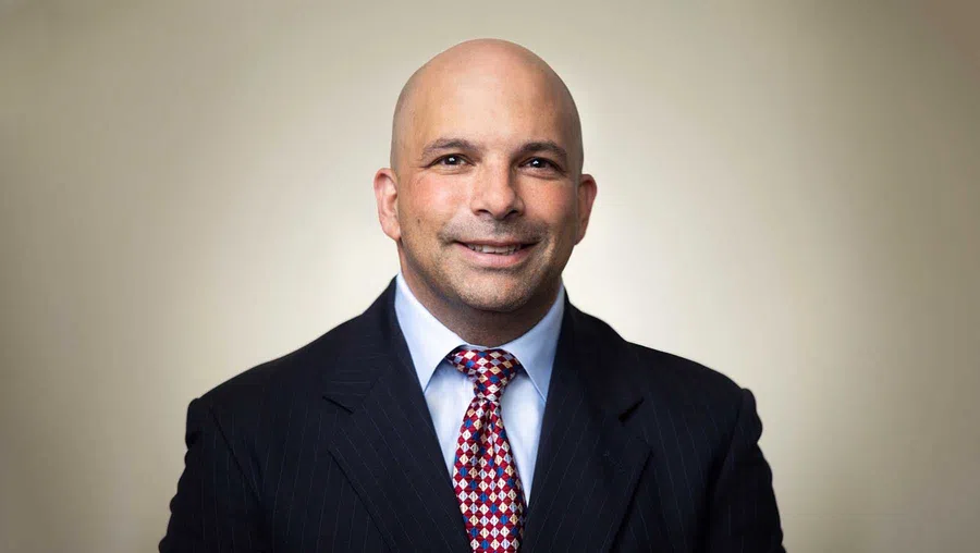 Smiling bald man in a dark suit with a red and white patterned tie against a neutral background.