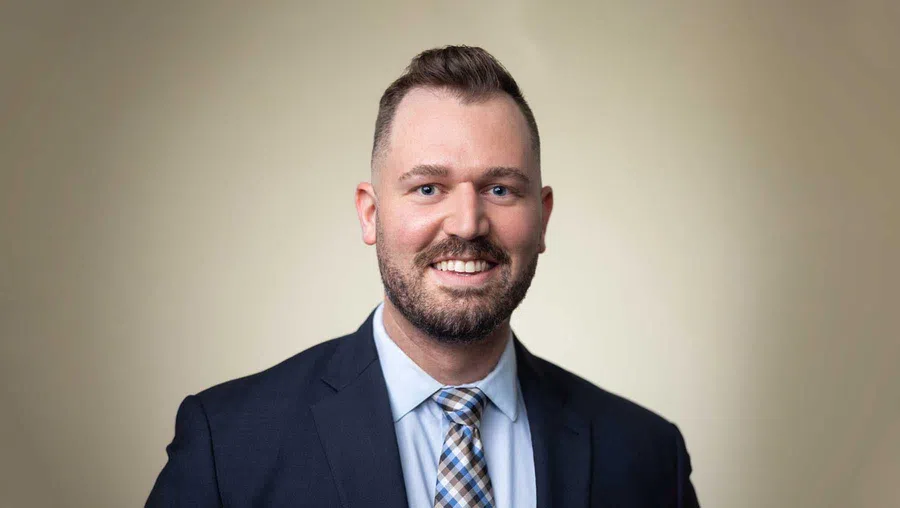 Smiling man with beard wearing a dark suit and patterned tie against a neutral background.