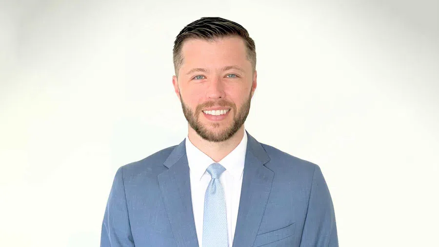 Smiling young man in blue suit and light blue tie against plain white background.