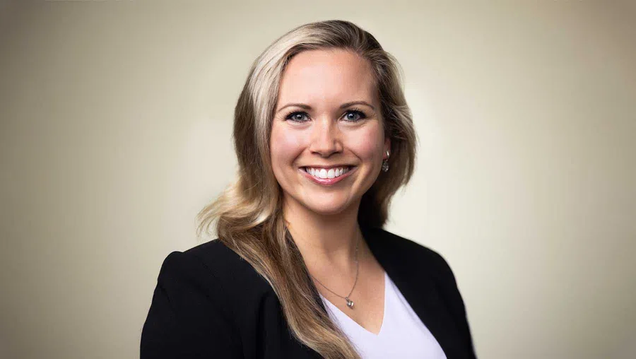 Smiling woman with long blonde hair wearing a black blazer and white top against a neutral background.