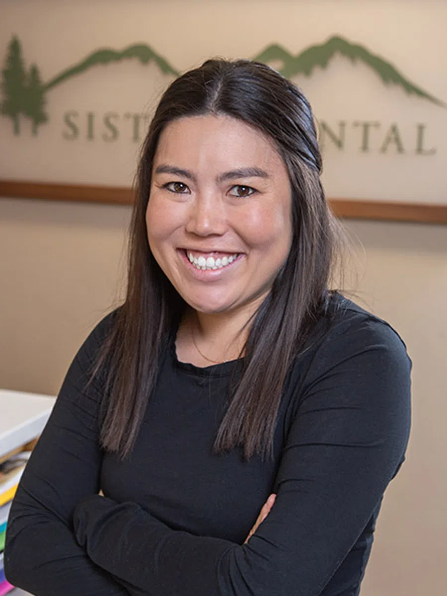 Smiling woman with dark hair in black shirt stands with arms crossed in front of dental office logo.