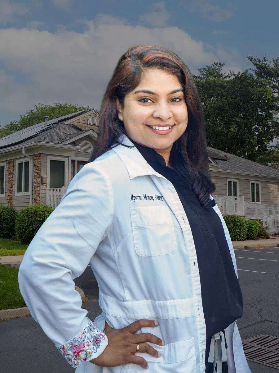 Smiling female dentist in white coat with Aparna Menon, DMD embroidered, in bright dental office.