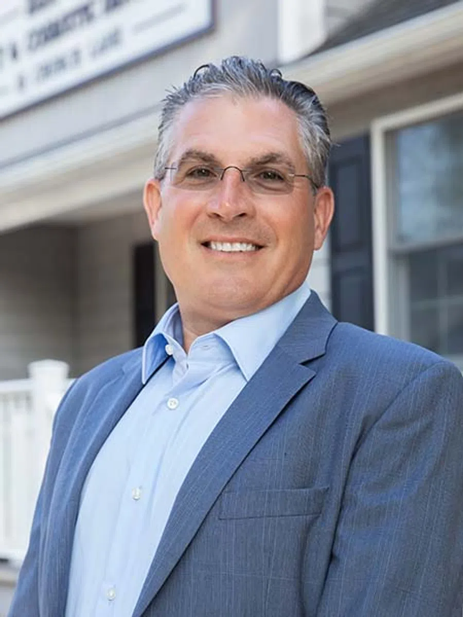 Smiling middle-aged man wearing glasses and a blue suit jacket standing outside a residential home.