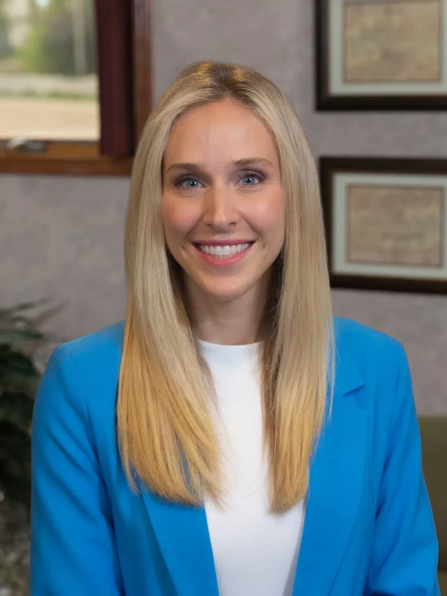 Smiling blonde woman in a blue blazer and white shirt posing indoors with framed pictures in the background.
