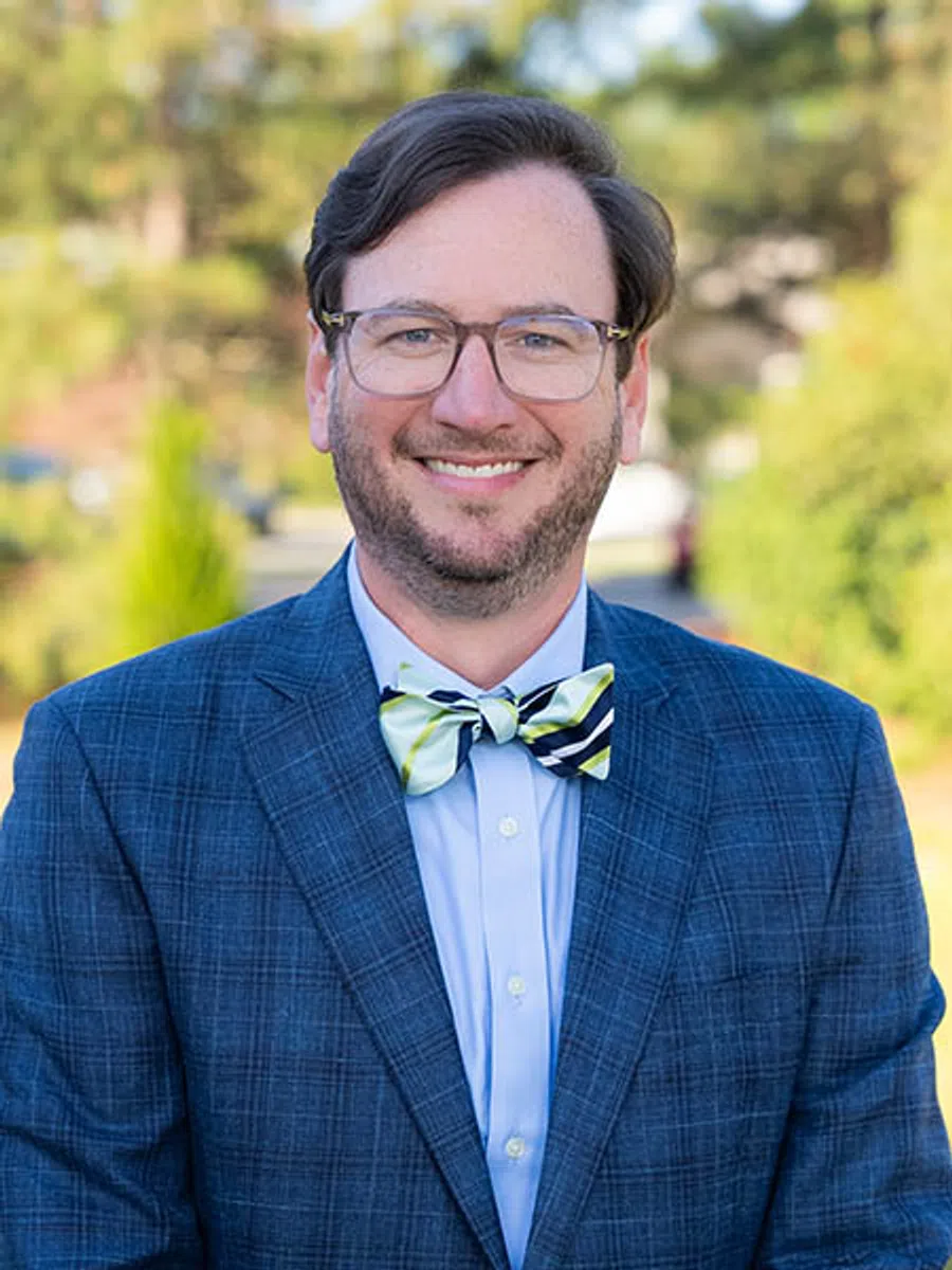 Smiling man wearing glasses, a blue checkered suit, light blue shirt, and a striped bow tie outdoors.