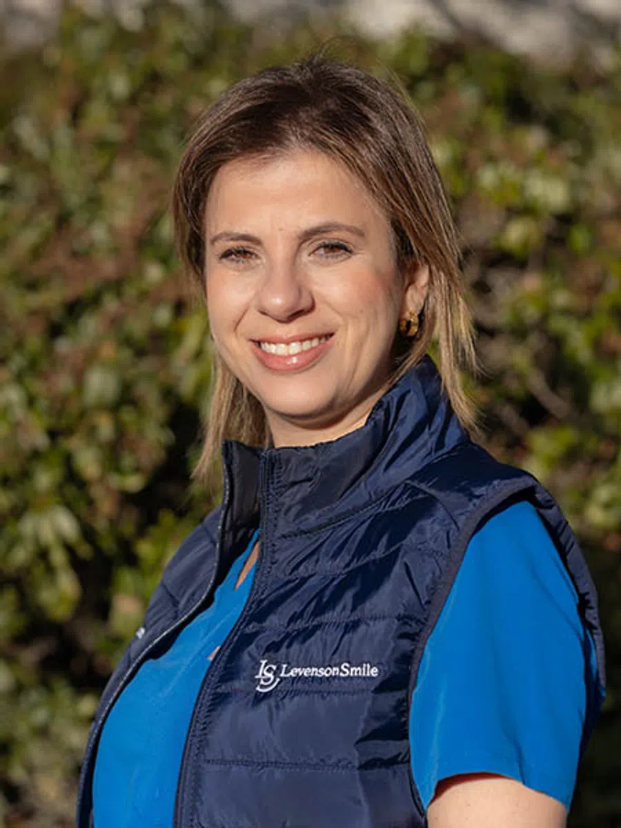 Smiling woman wearing a blue medical scrubs and navy vest with Levenson Smile logo outside in sunlight.