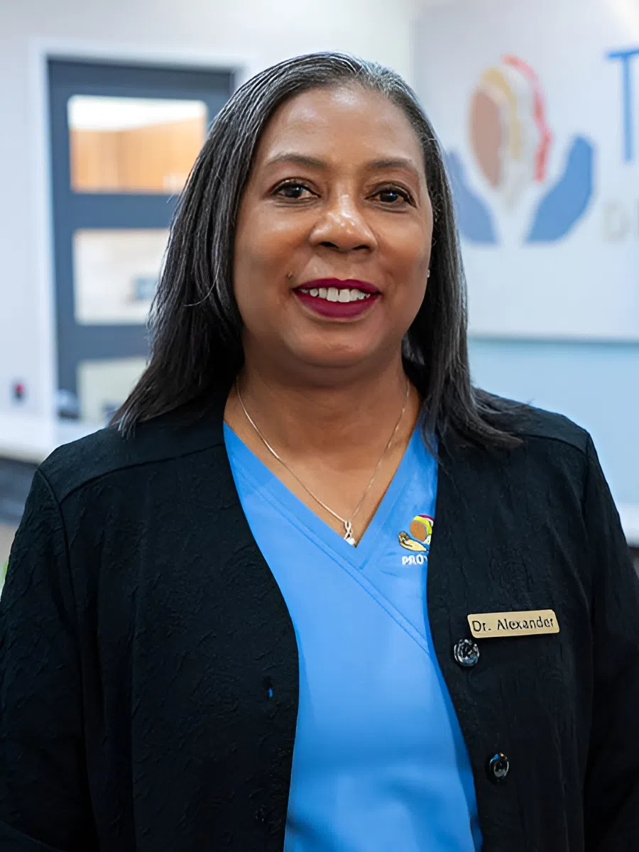 Smiling female doctor wearing a name tag and blue scrubs standing in a medical office environment.