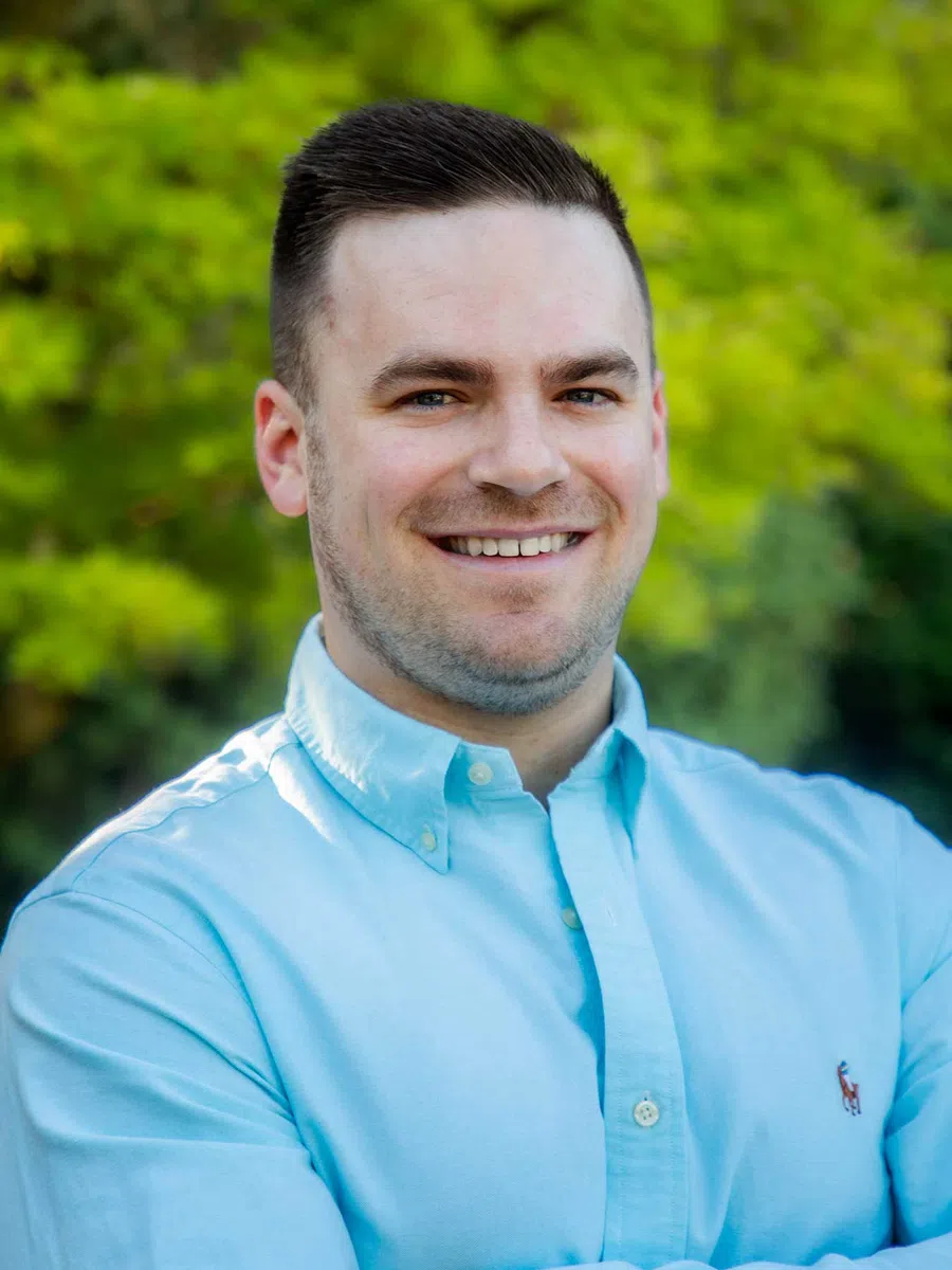 Smiling man in light blue button-up shirt posing outdoors with green trees in the background.