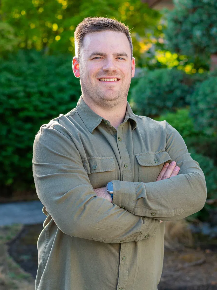 Smiling man in green button-up shirt with arms crossed outdoors in a sunny garden setting.