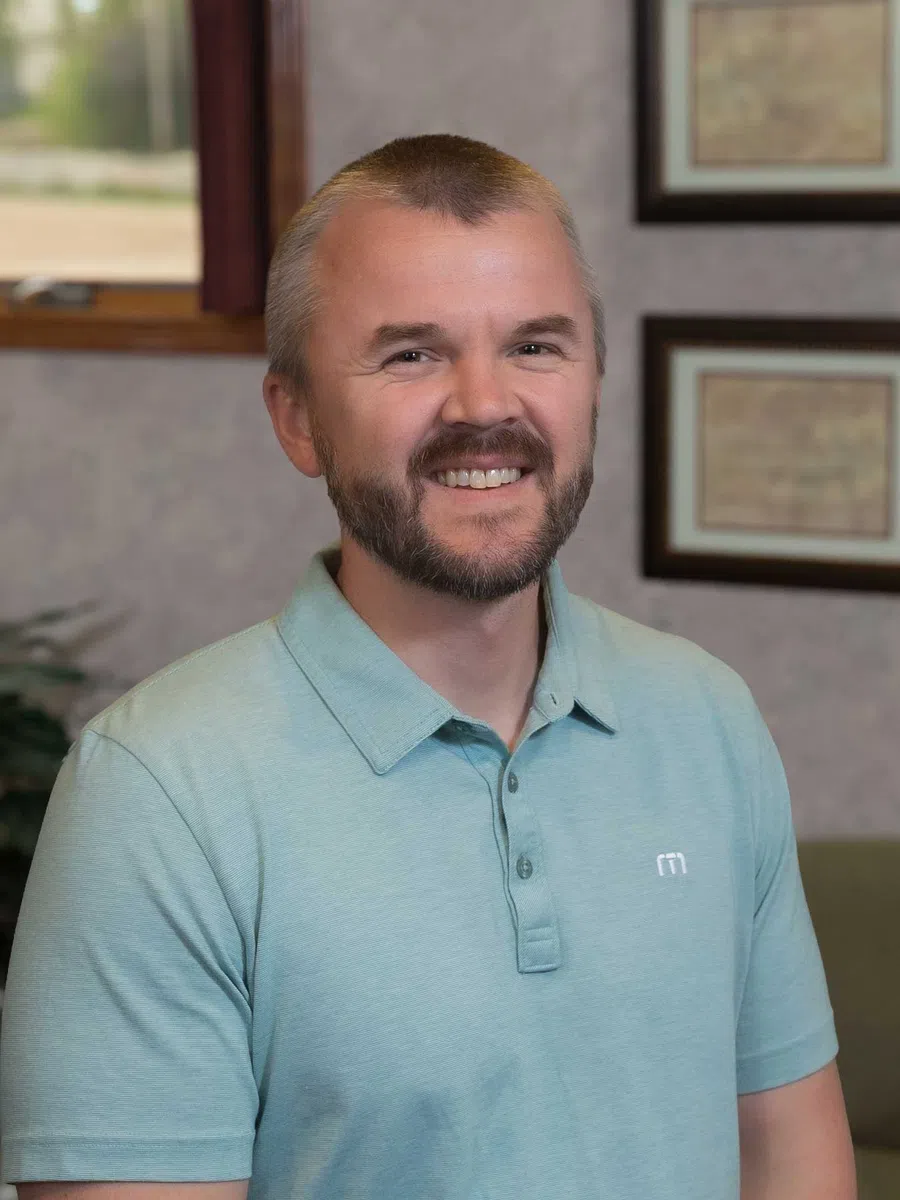 Smiling man with short beard and light blue polo shirt in a cozy indoor setting with framed pictures on the wall.