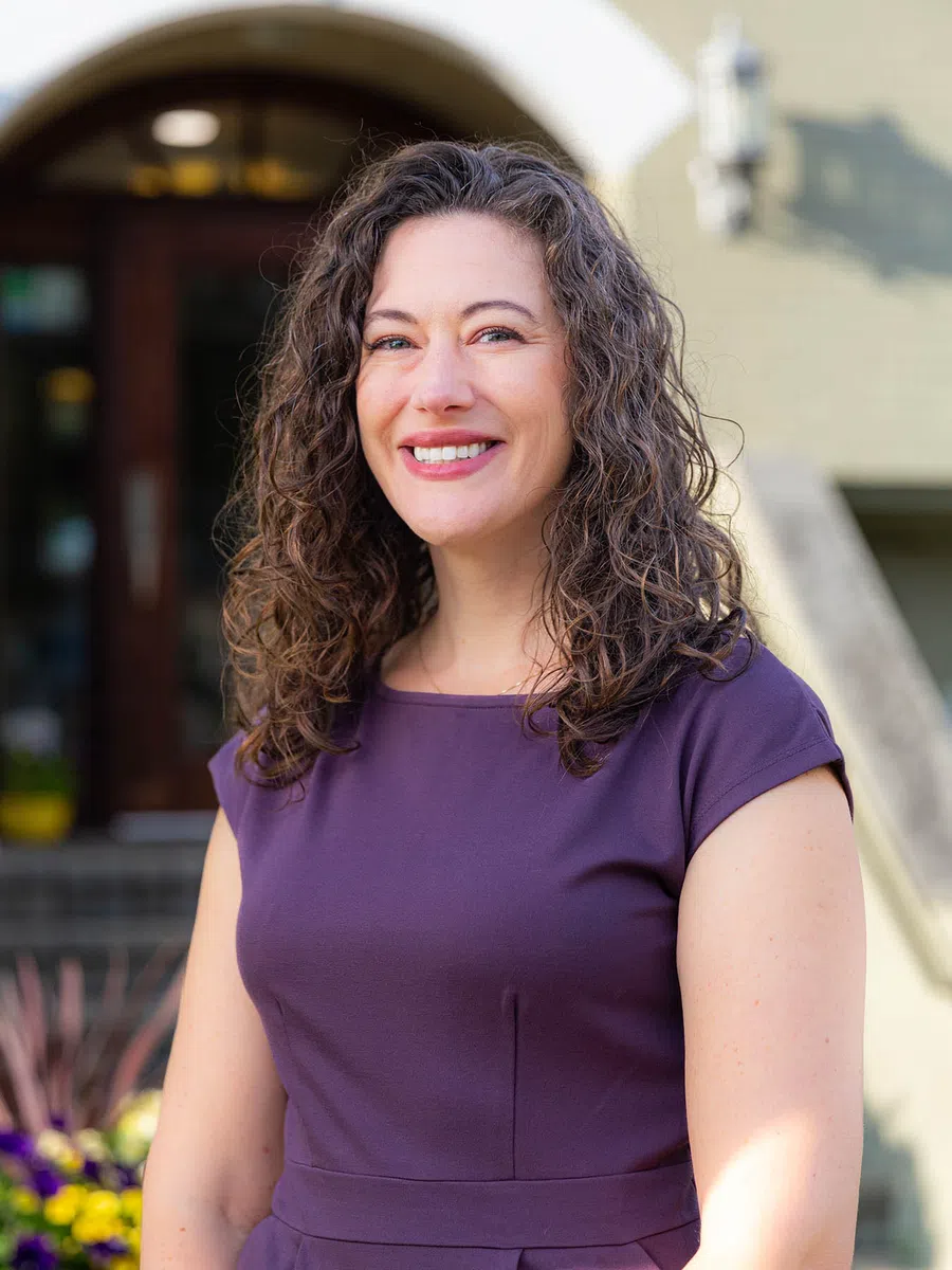 Smiling woman with curly brown hair wearing a purple dress standing outdoors in front of a building.