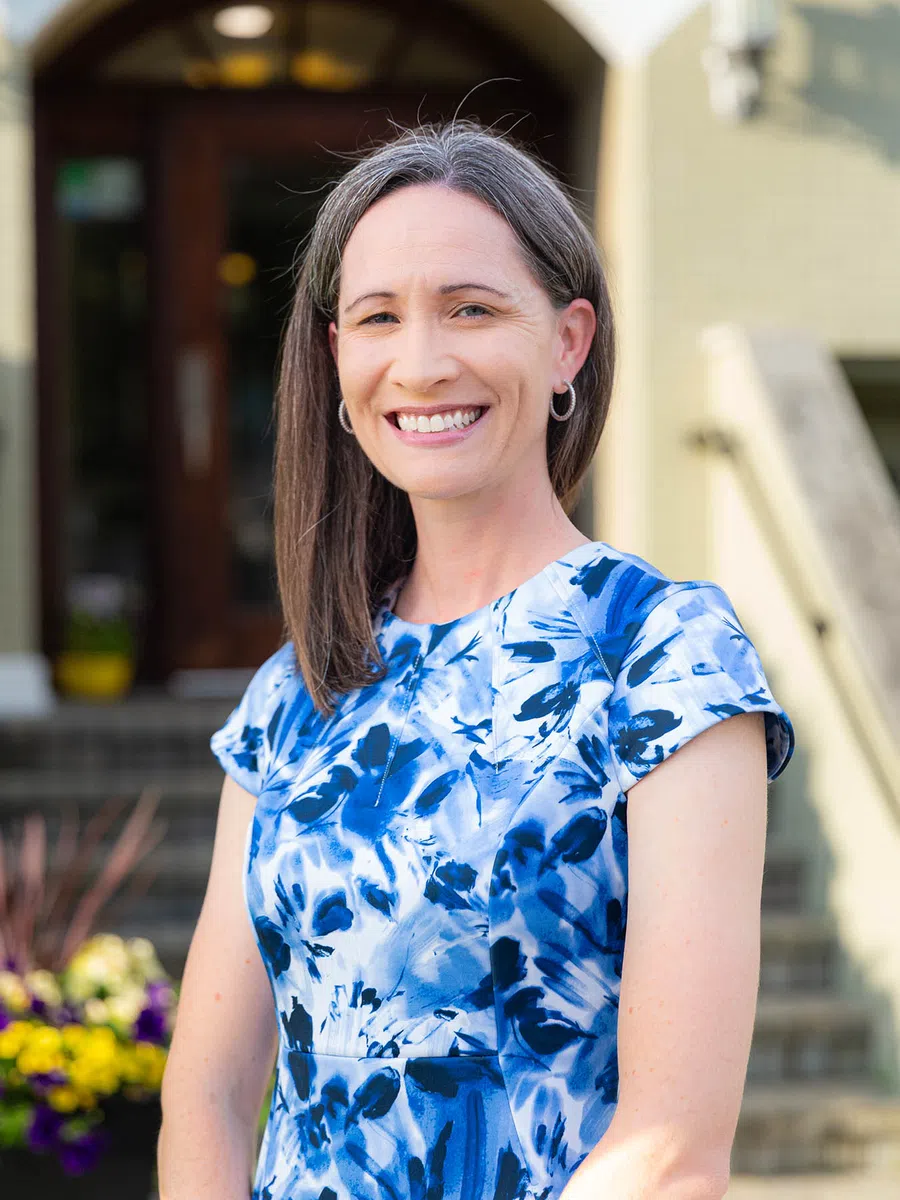 Smiling woman with straight brown hair wearing a blue and white floral dress standing outdoors near stairs.