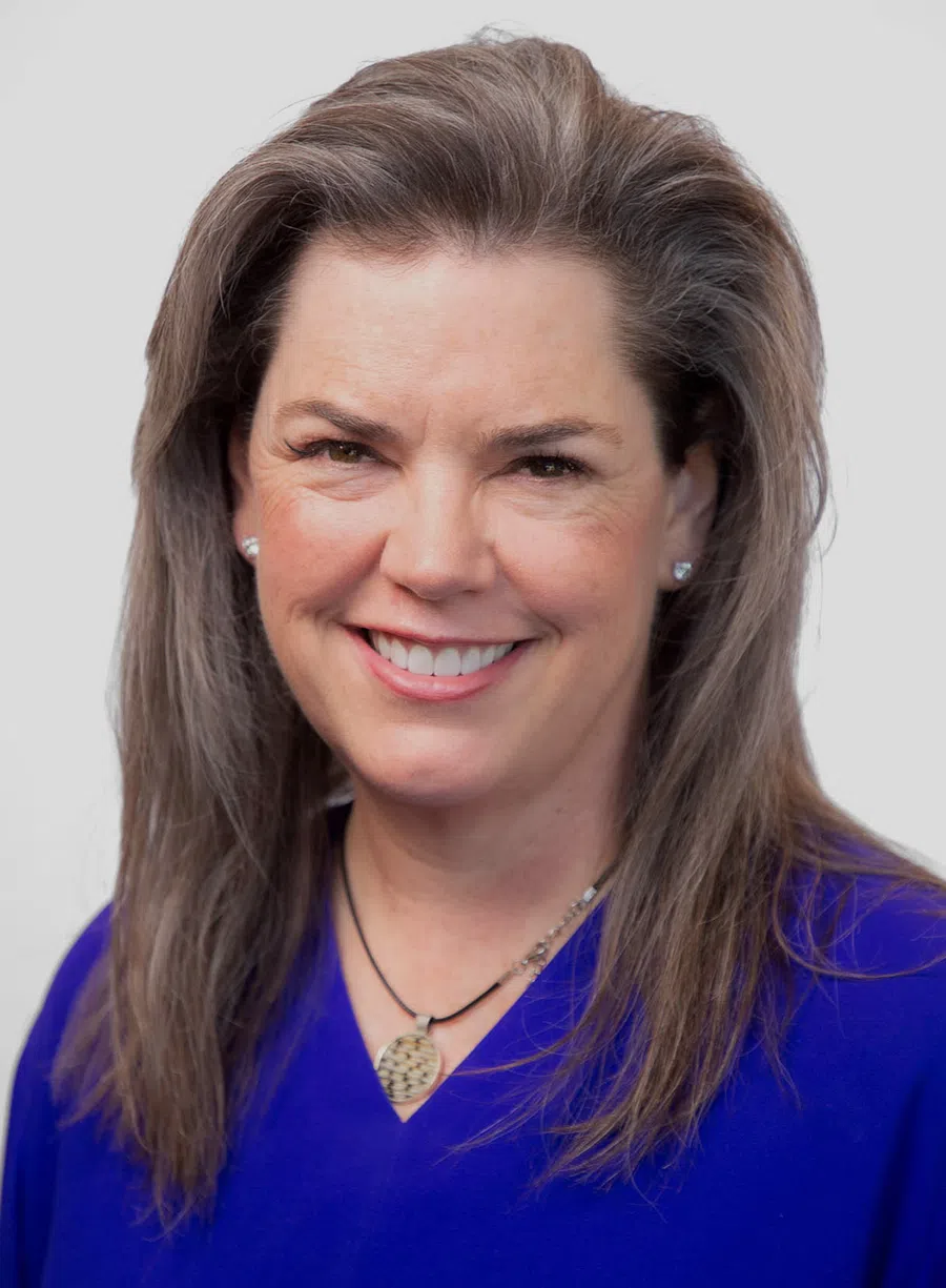 Smiling middle-aged woman with gray hair wearing a blue top and heart-shaped earrings against a plain background