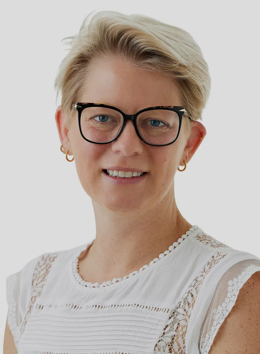 Blonde woman with short hair wearing tortoiseshell glasses and white lace-trimmed top smiling at camera.